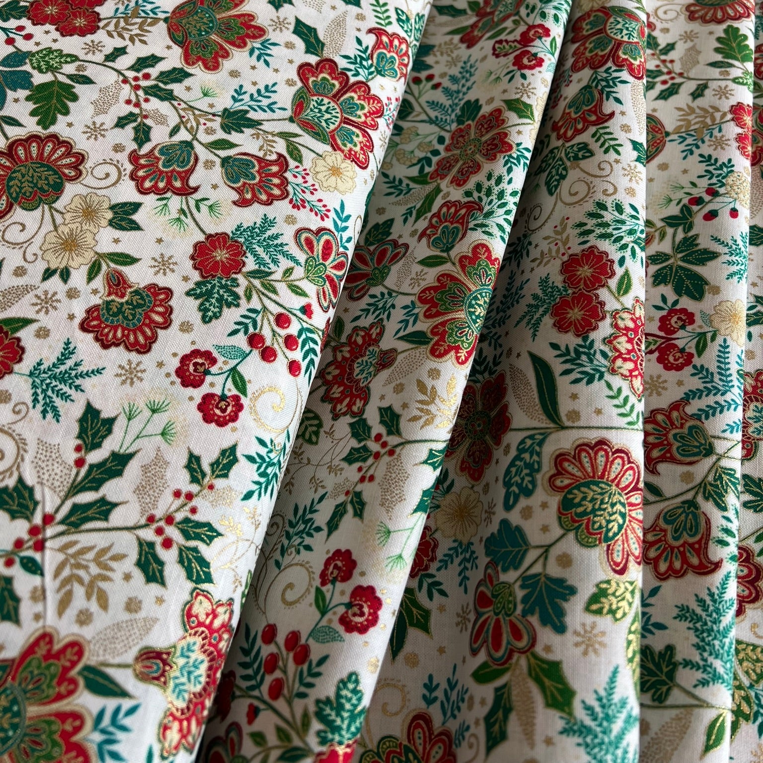 A close-up of draped fabric with a dense floral and botanical print on an off-white background, featuring red, green, gold metallic and beige flowers and leaves.