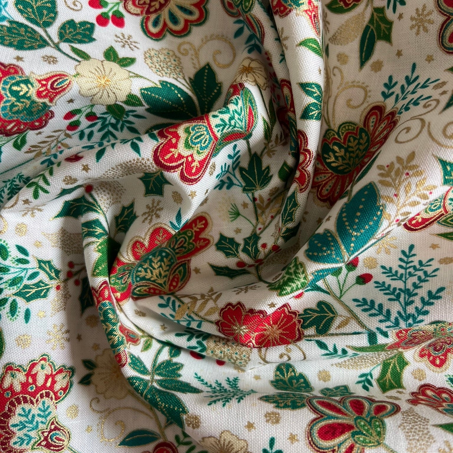 A close-up of draped fabric with an all-over floral and botanical print on a warm off-white background. The pattern features red and green flowers, green leaves, and small gold decorative swirls.