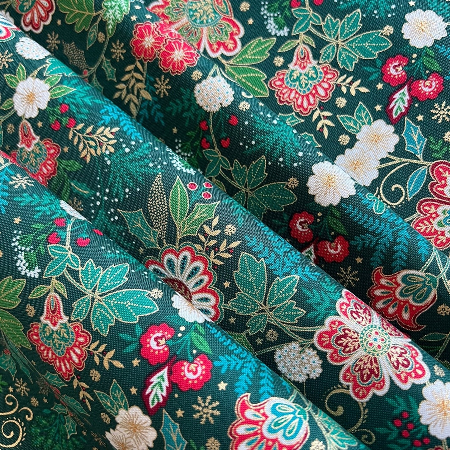 A close-up of a folded fabric with a dense floral and botanical print on a dark teal-green background. The pattern features red, pink, and white flowers, light green leaves, and small gold star-like accents.