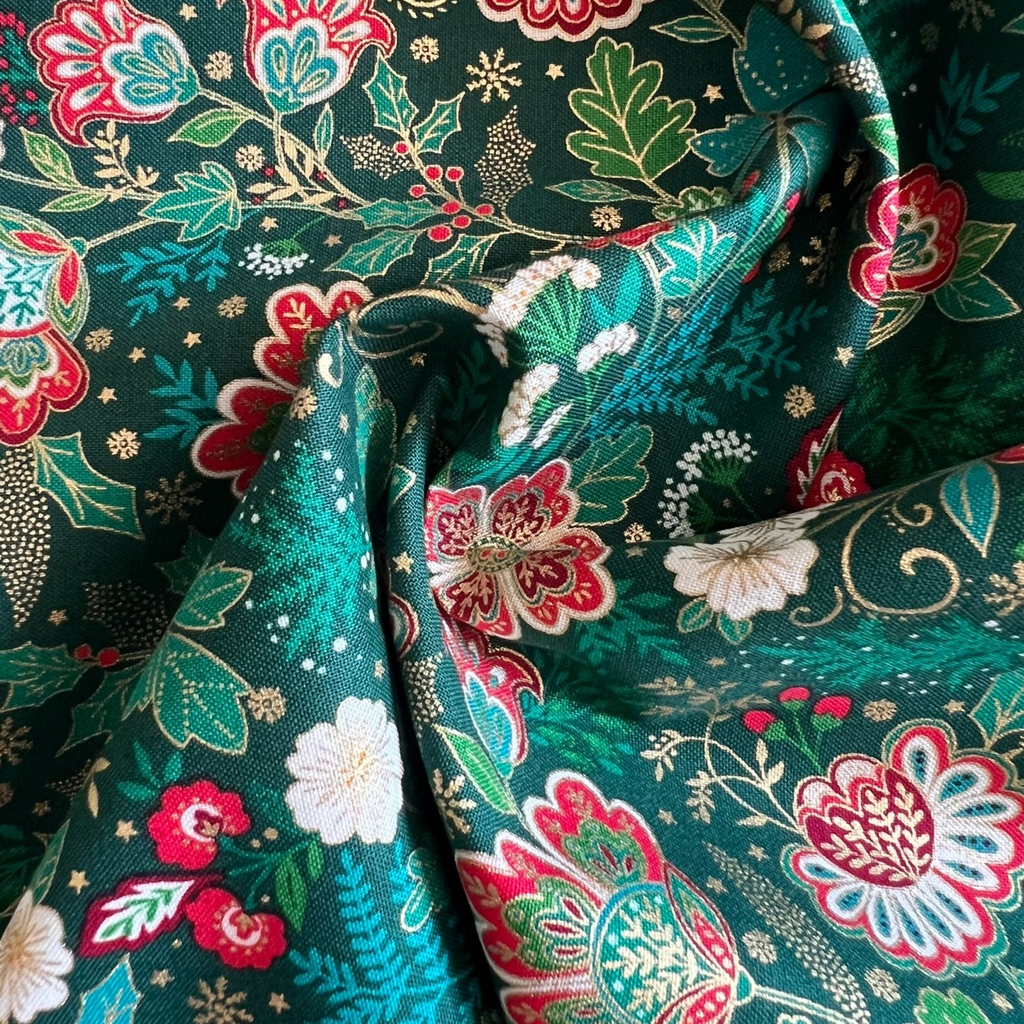A close-up of draped fabric with an intricate floral and botanical print on a dark teal-green background. The pattern features red, pink, white, and light green flowers with gold outlines and accents.