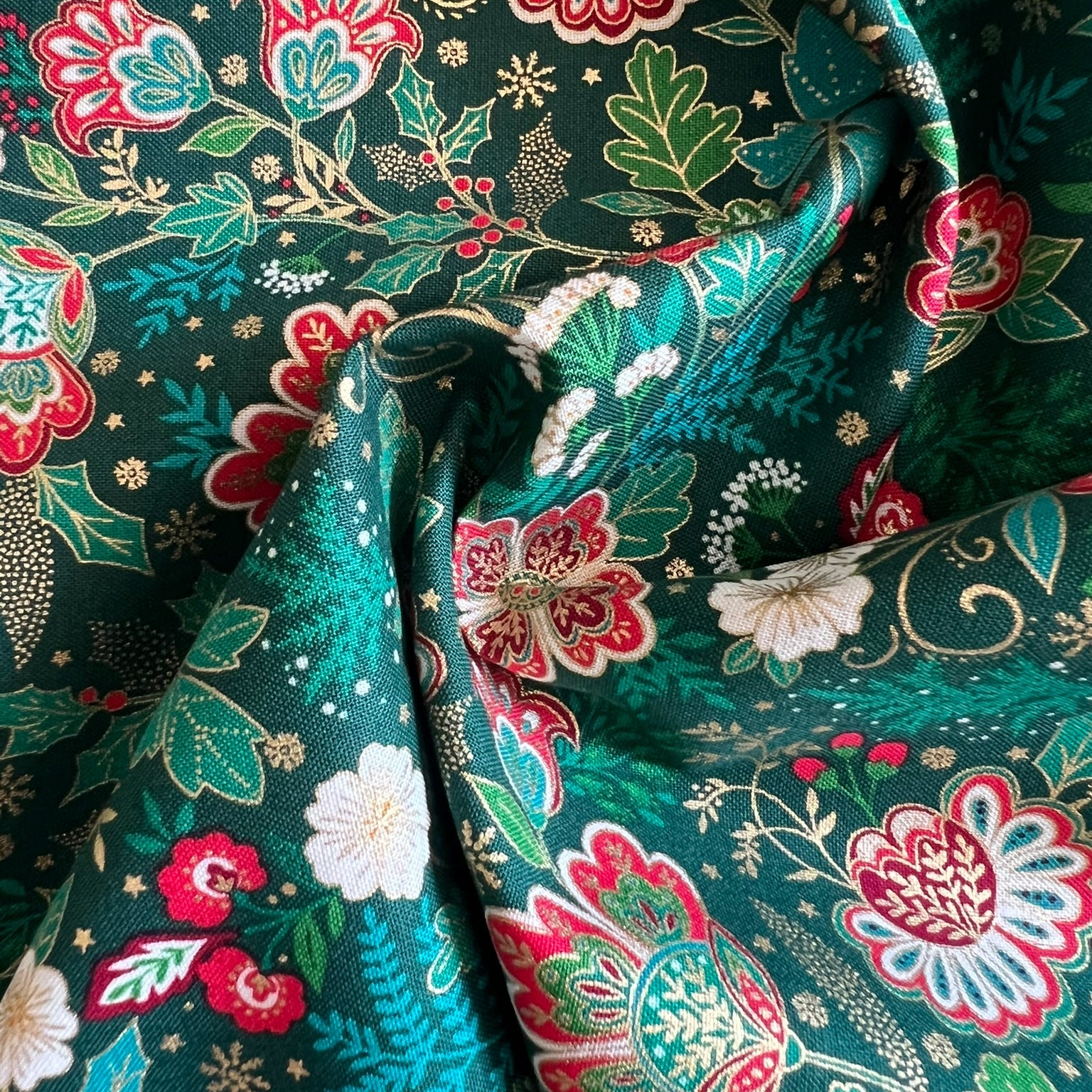 A close-up of draped fabric with an intricate floral and botanical print on a dark teal-green background. The pattern features red, pink, white, and light green flowers with gold outlines and accents.