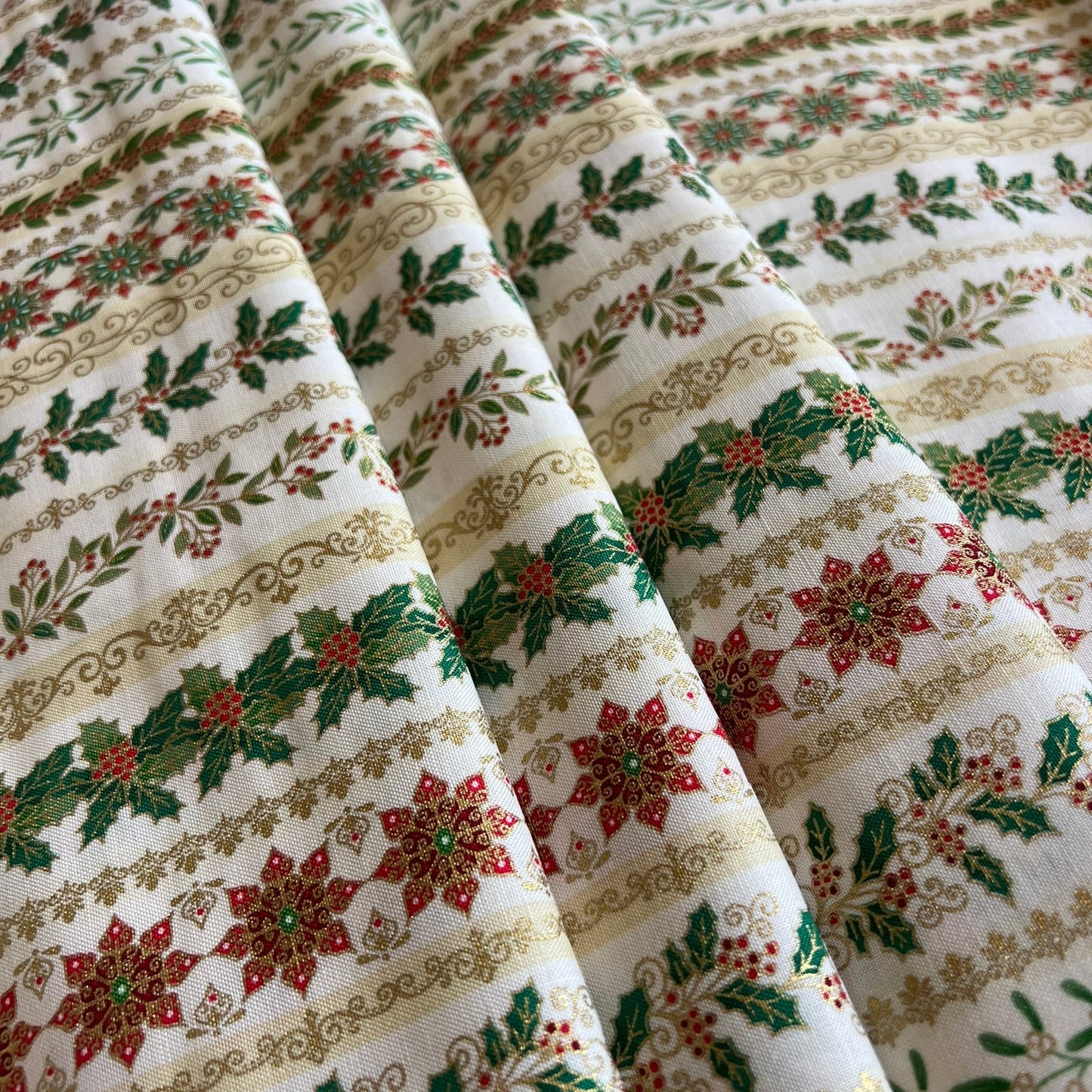 A close-up of a draped fabric with a festive holiday pattern featuring red poinsettias, green holly leaves, and gold metallic decorative borders on an off-white background.