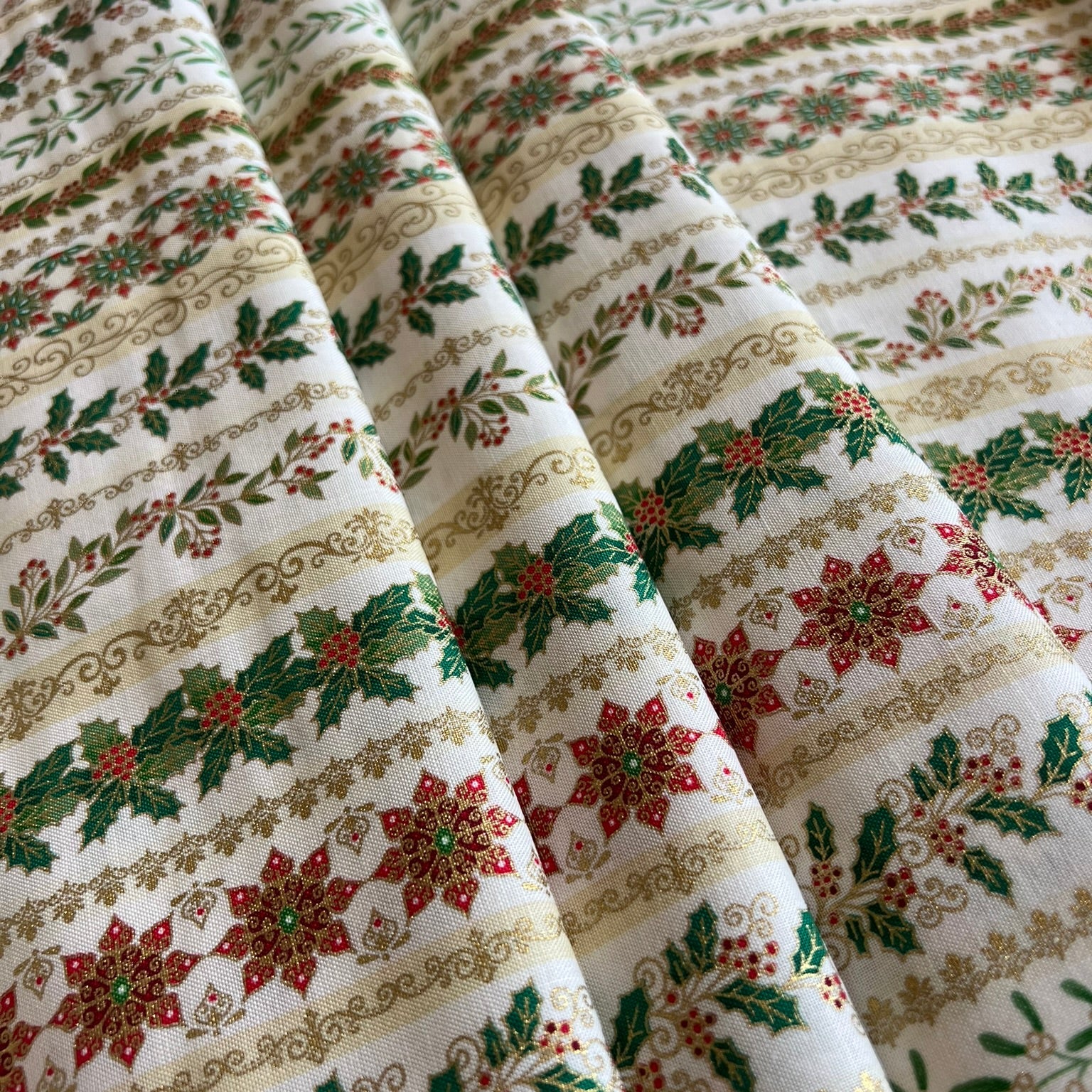 A close-up of a draped fabric with a festive holiday pattern featuring red poinsettias, green holly leaves, and gold metallic decorative borders on an off-white background.