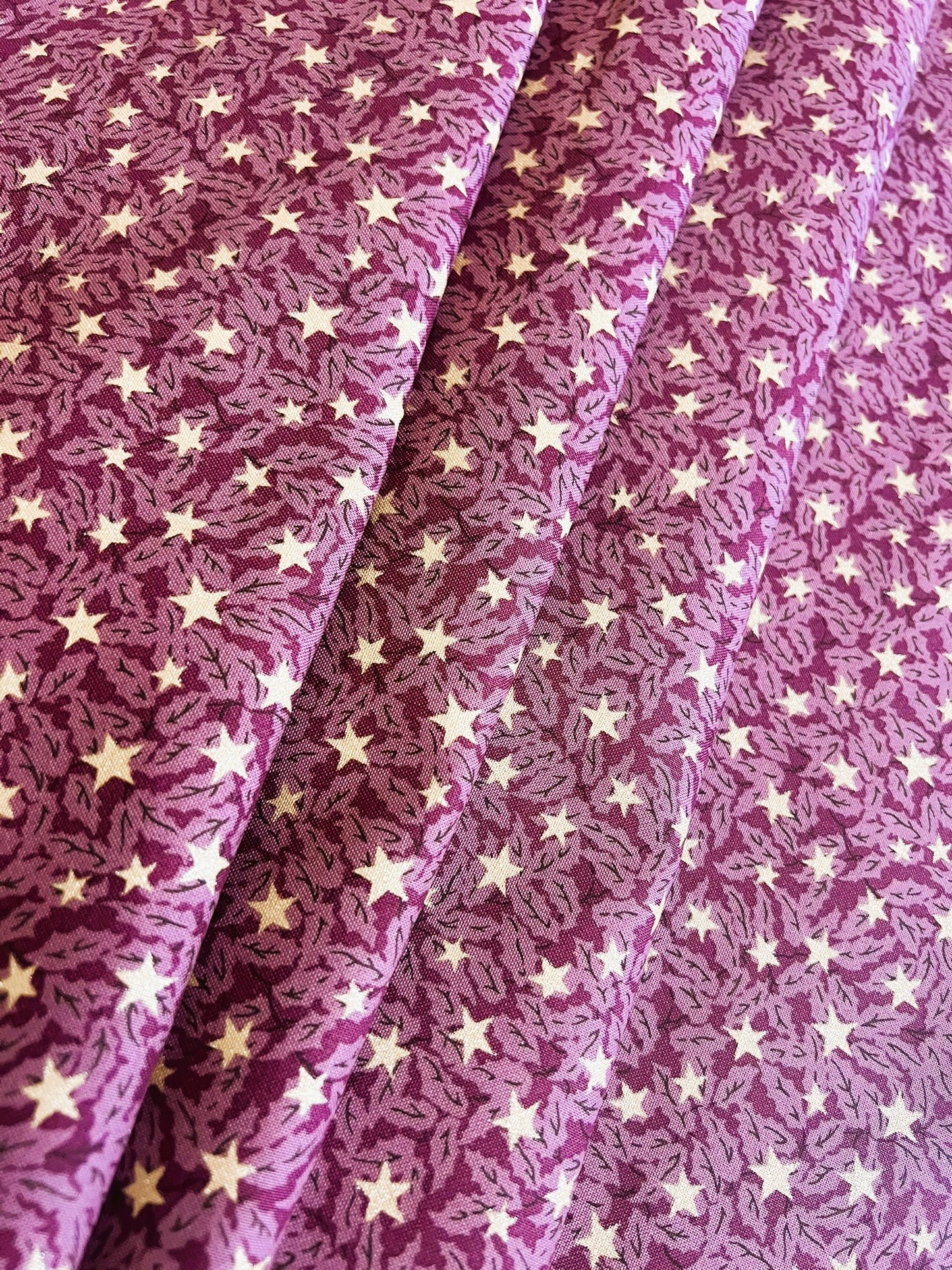 A close-up of a purple fabric with a repeating pattern of small cream stars and fine wavy lines, shown in soft folds.