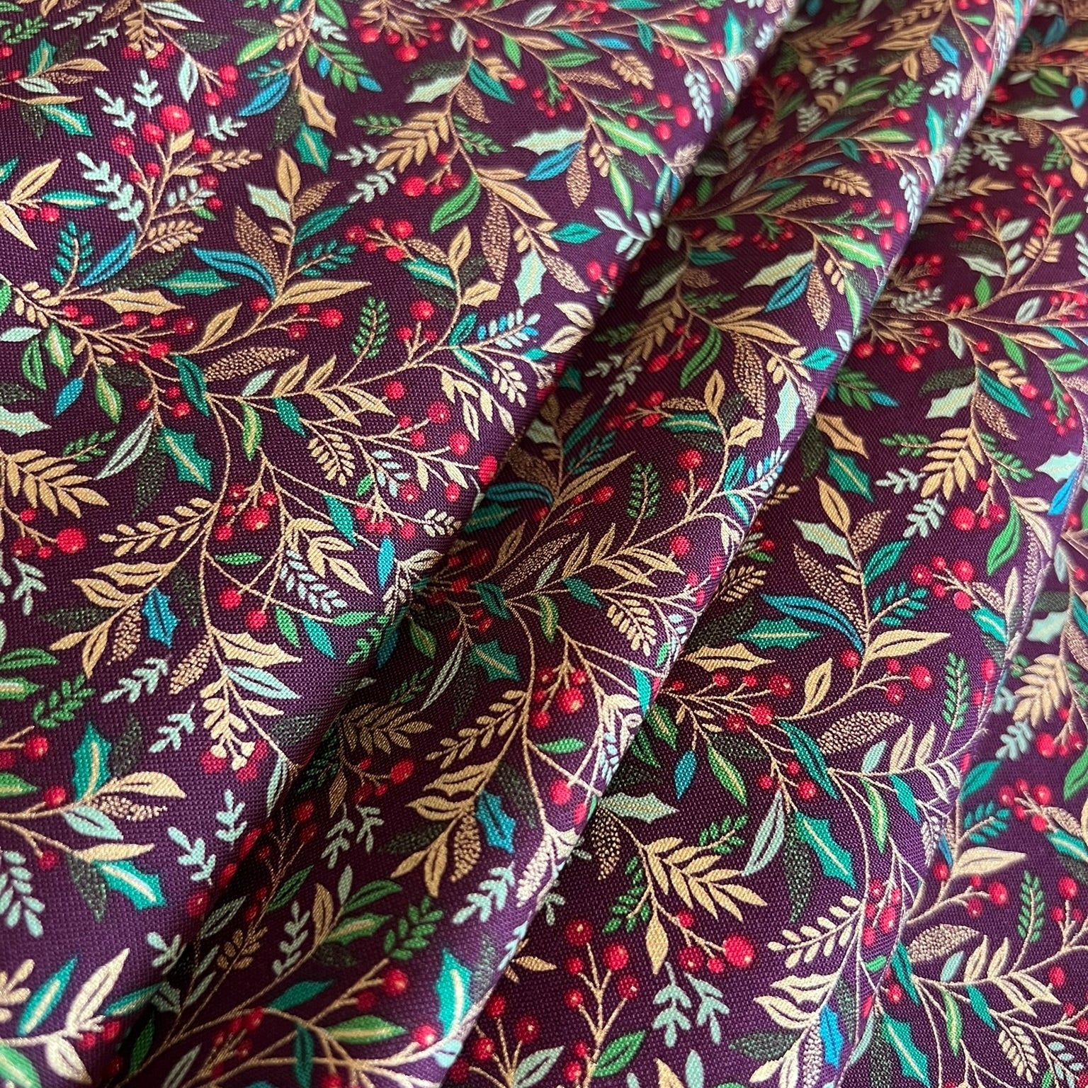 A close-up of draped fabric with an all-over botanical print featuring burgundy berries, green and metallic gold leaves, and tan foliage on a dark burgundy background.