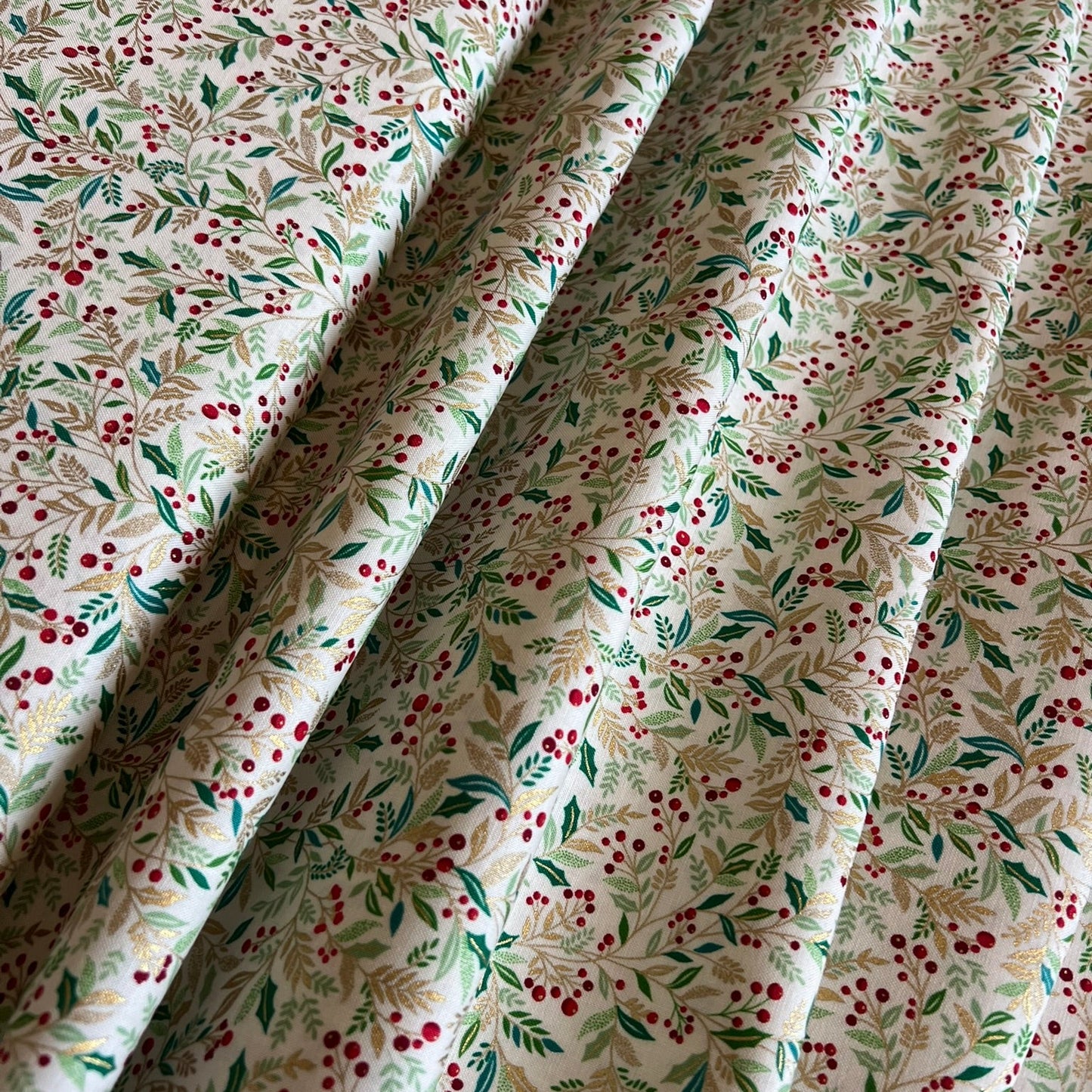 A close-up of draped fabric with a delicate botanical print featuring gold metallic and green leaves and small red berries on an off-white background.