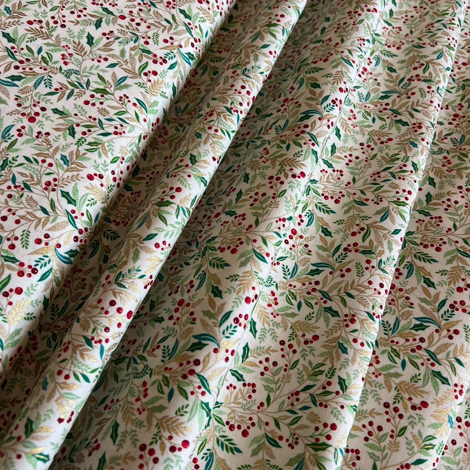 A close-up of draped fabric with a delicate botanical print featuring gold metallic and green leaves and small red berries on an off-white background.