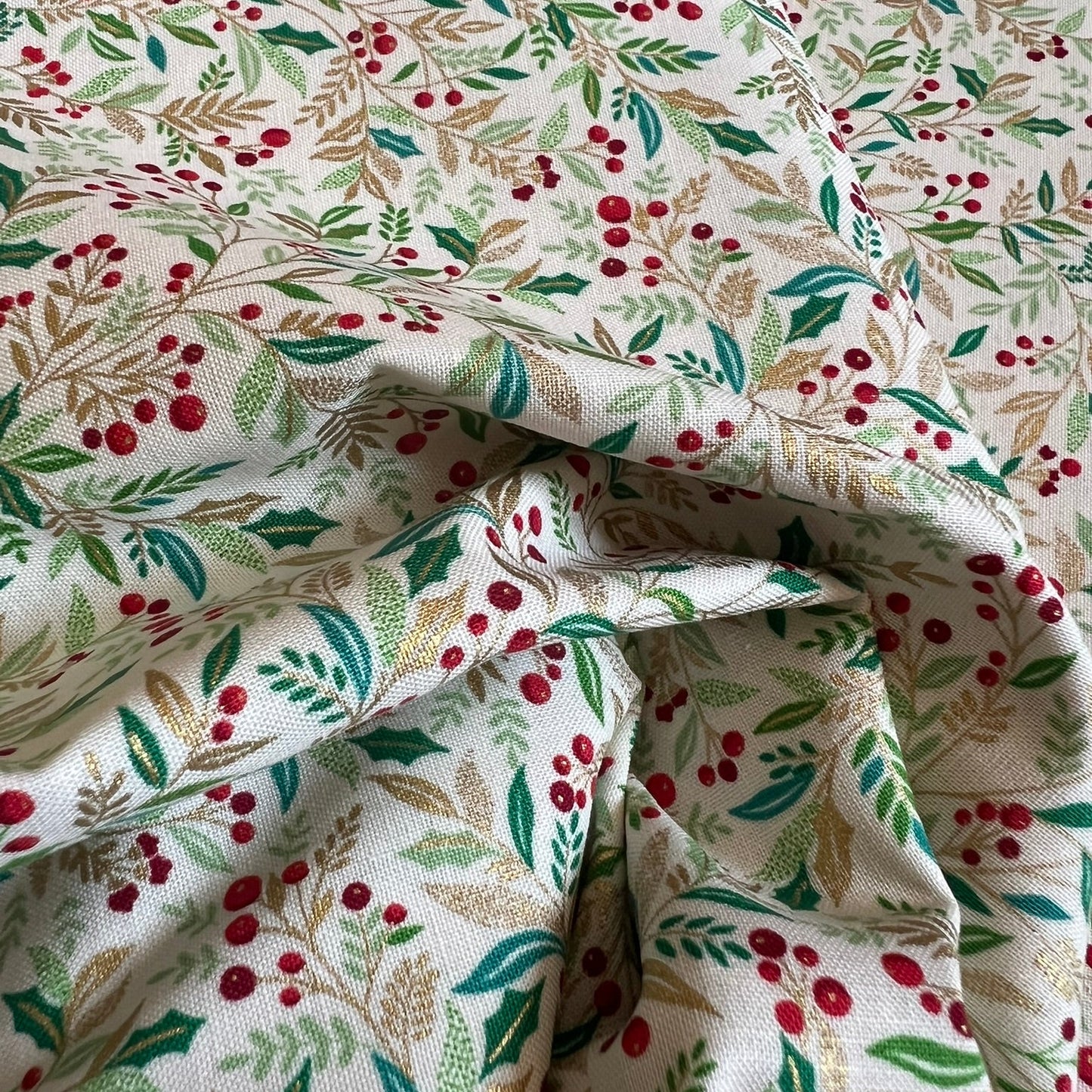 A close-up of draped fabric with an all-over botanical print featuring gold metallic and green leaves, tan stems, and small red berries on an off-white background.