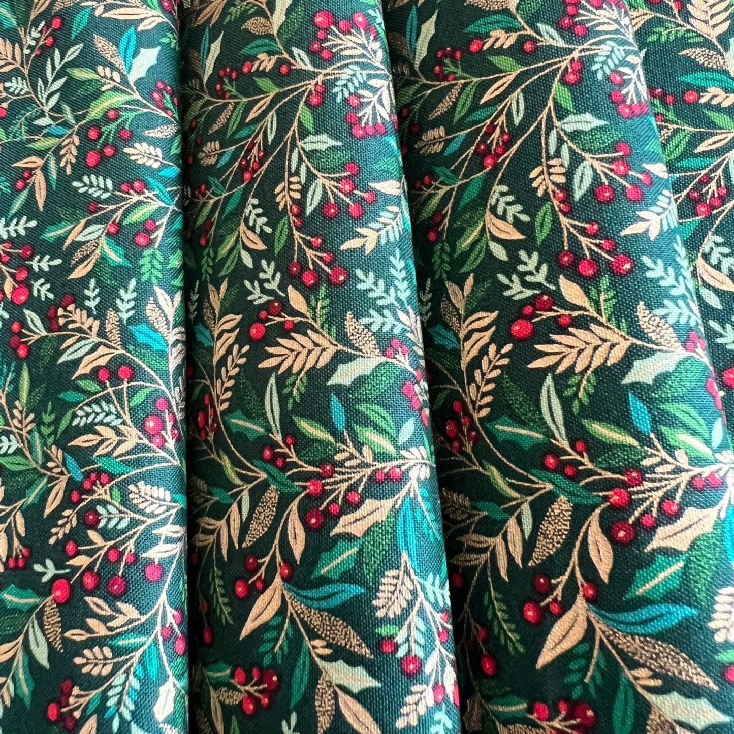 A close-up of draped fabric with an all-over botanical print featuring green and gold metallic leaves, beige branches, and clusters of small red berries on a dark teal-green background.