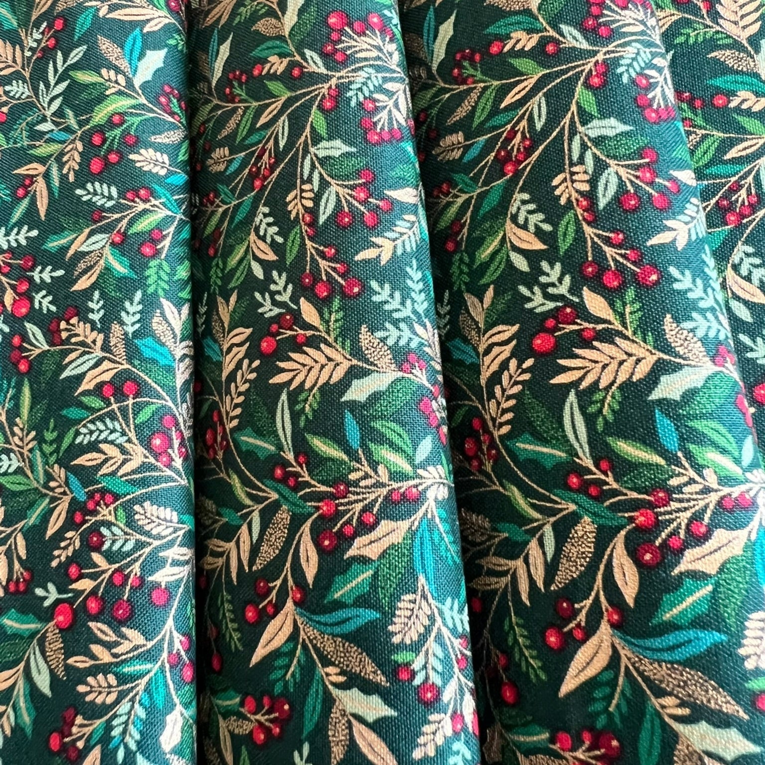 A close-up of draped fabric with an all-over botanical print featuring green and gold metallic leaves, beige branches, and clusters of small red berries on a dark teal-green background.