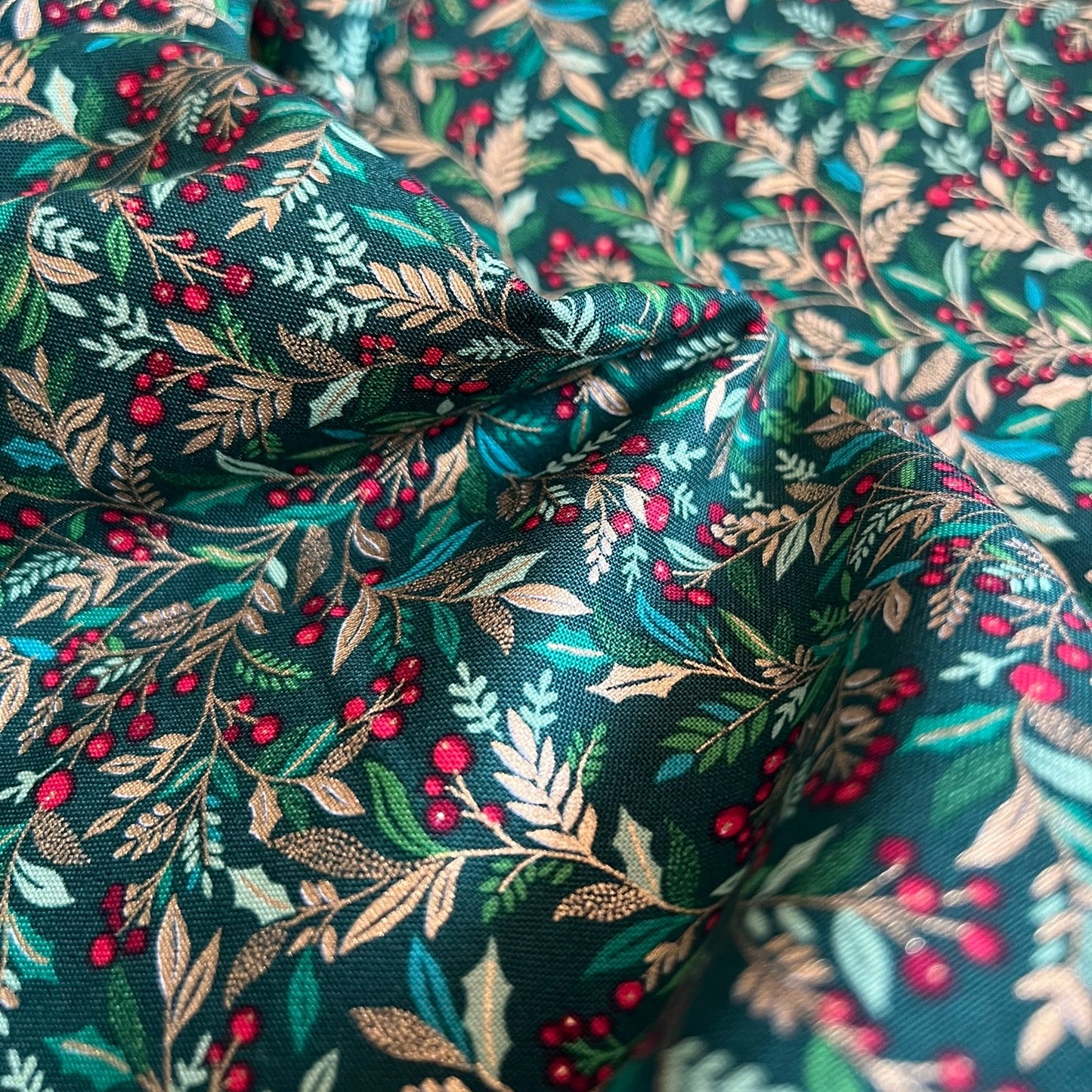 A close-up of draped fabric with an all-over botanical print featuring green and metallic gold leaves, beige branches, and clusters of small red berries on a dark teal background.