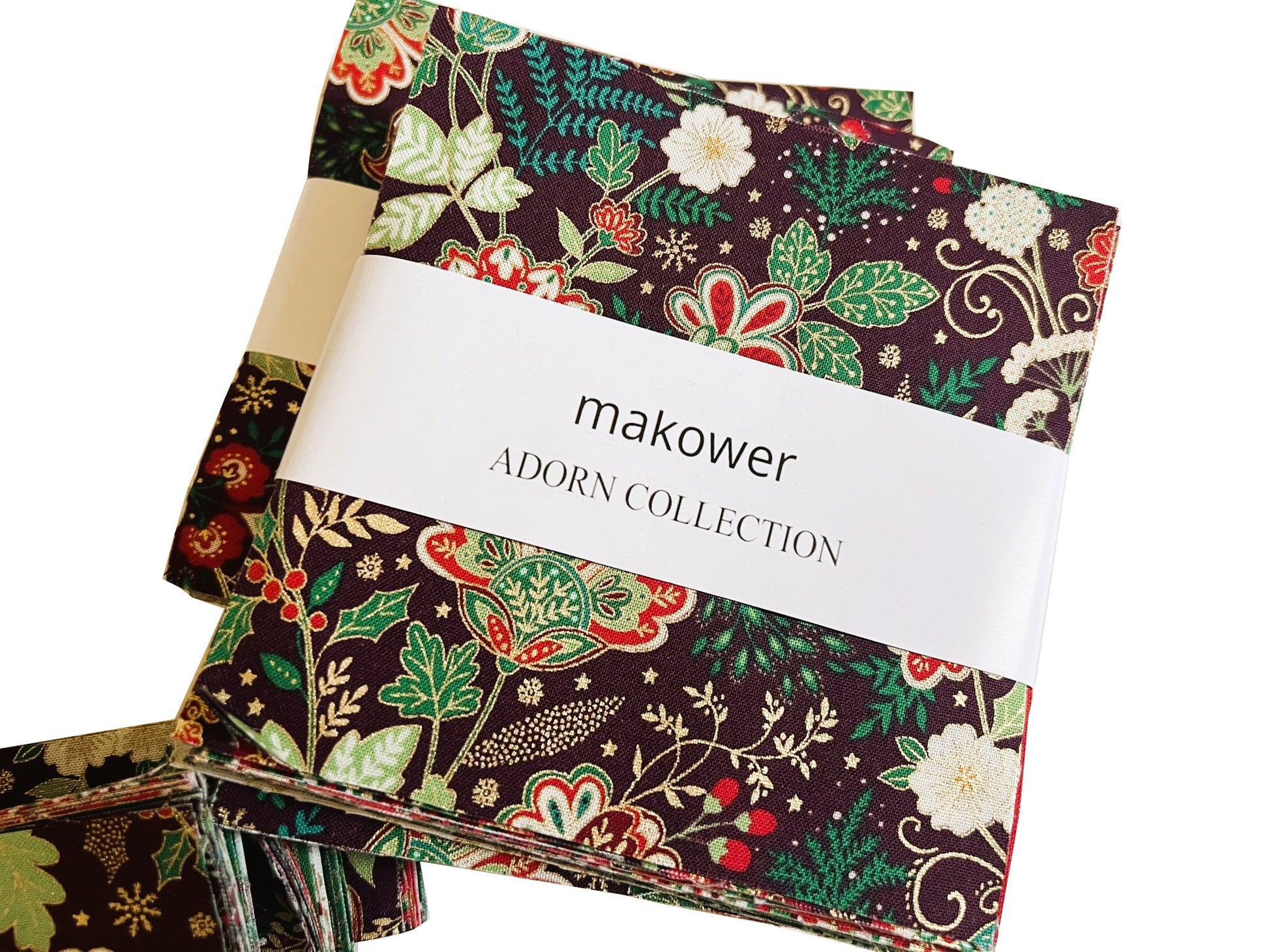 A stack of fabric swatches from the makower Adorn Collection, featuring a dark brown background with a detailed botanical pattern in green, red, and cream, wrapped with a white paper band.