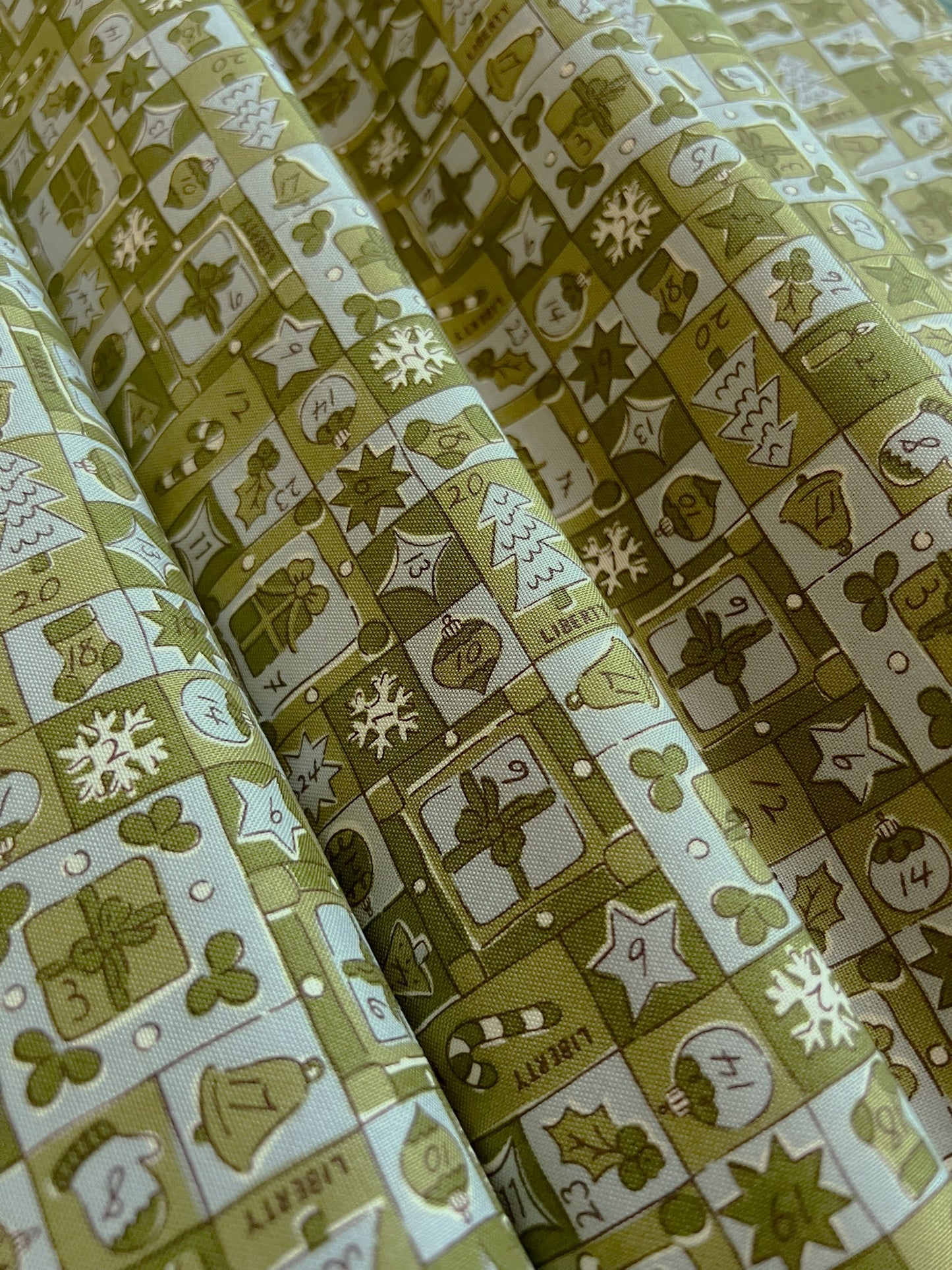 A close-up of a patterned fabric with a warm olive green and cream color scheme, featuring a grid of holiday-themed squares with illustrations of snowflakes, ornaments, stars, and the word 'Liberty'.