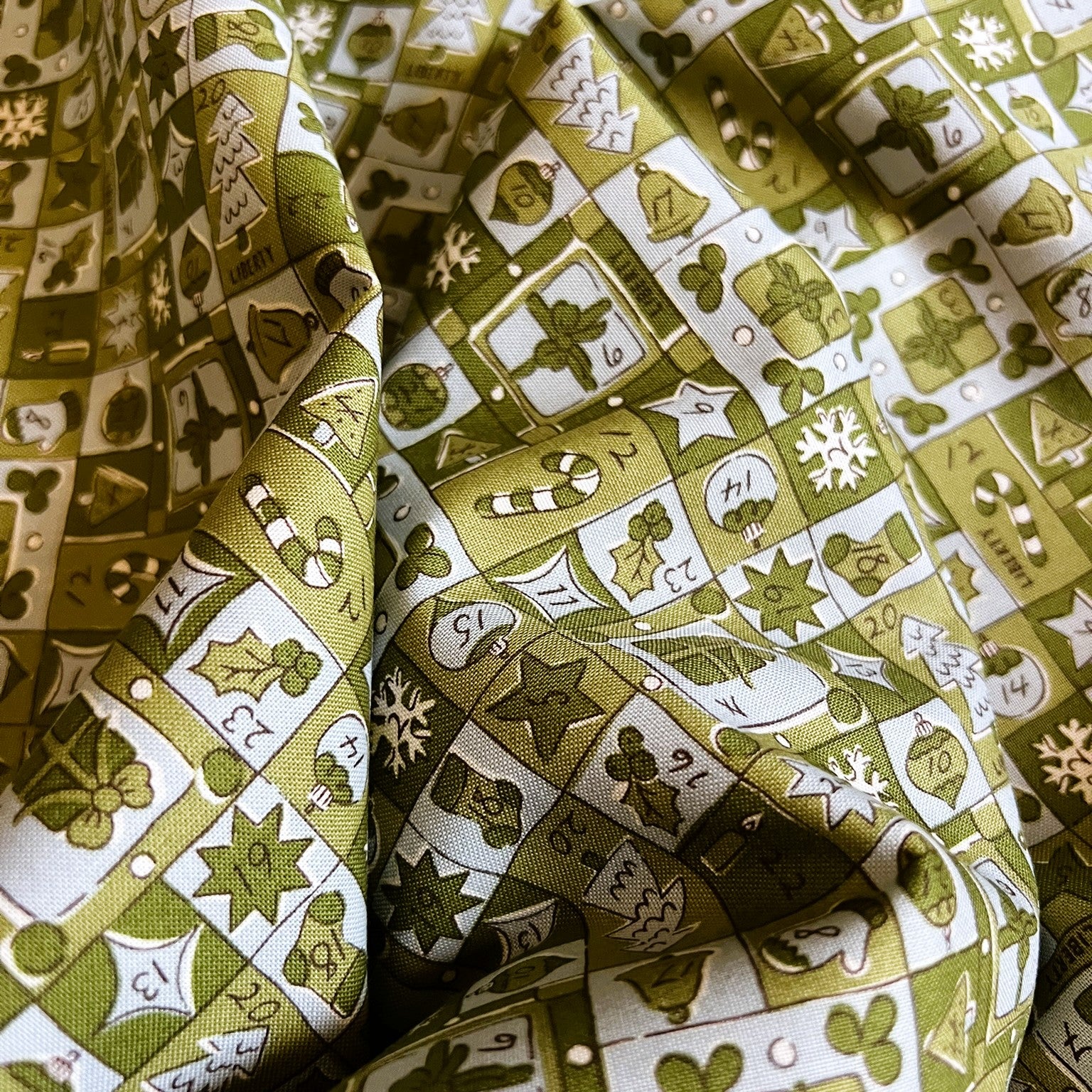 A close-up of draped fabric with an all-over holiday-themed print in olive green, cream, and white, featuring illustrations of candy canes, snowflakes, clovers, stars, and ornaments.