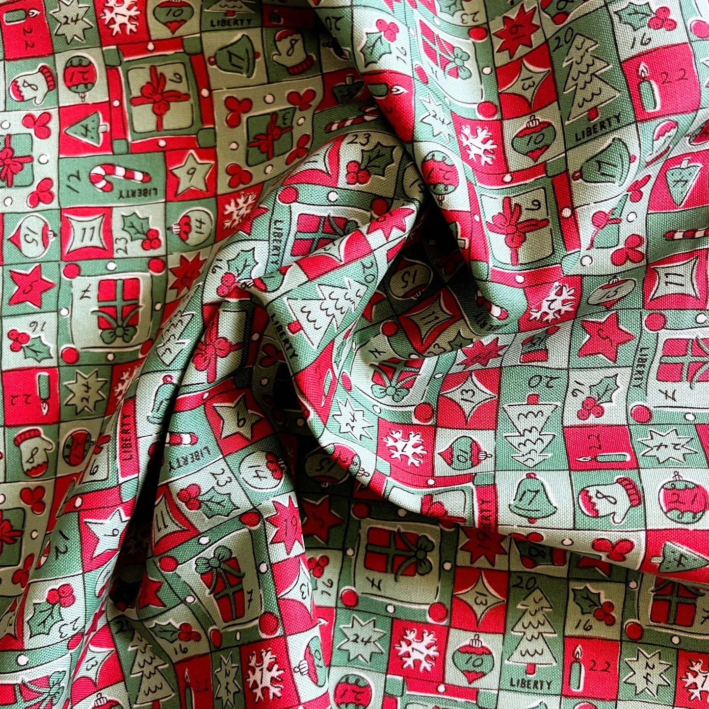 A close-up of draped holiday-themed fabric with a repeating pattern of red, green, and cream Christmas motifs including gifts, trees, candy canes, stars, and ornaments.