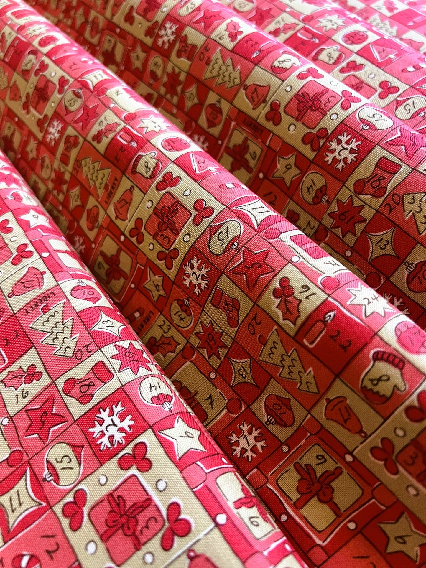 Red and beige patterned fabric with a holiday grid design featuring stockings, gifts, snowflakes, and stars, shown in soft folds.
