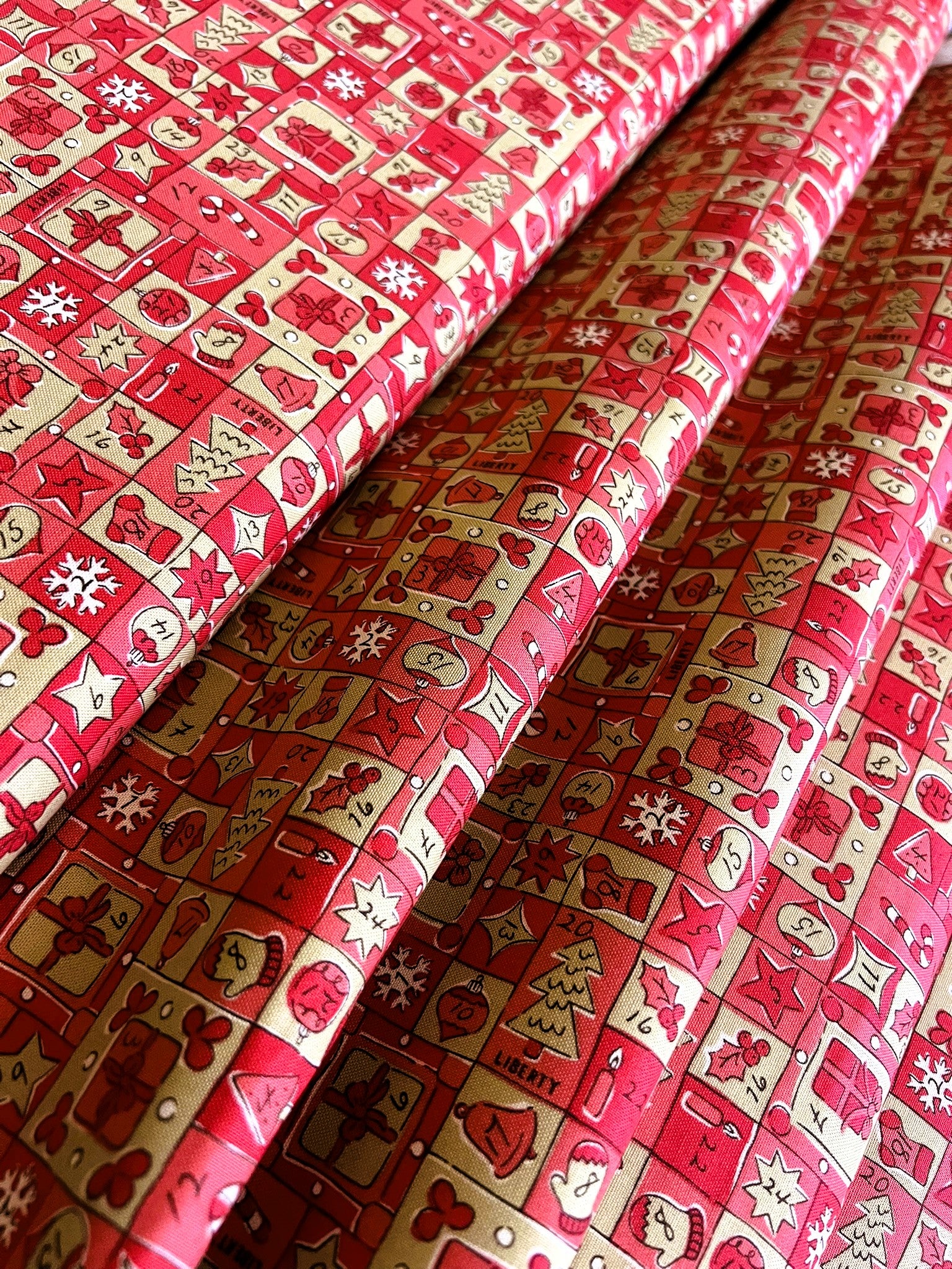 Red and gold patterned fabric with a festive holiday design featuring gingerbread men, snowflakes, ornaments, and gift boxes, shown in soft folds.