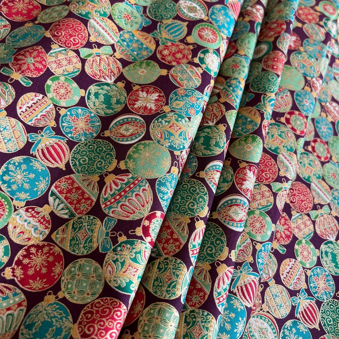 A close-up of patterned fabric with a repeating Christmas ornament print on a dark burgundy background. The ornaments are in green, red, pink, and metallic gold, with some featuring snowflakes and striped details.