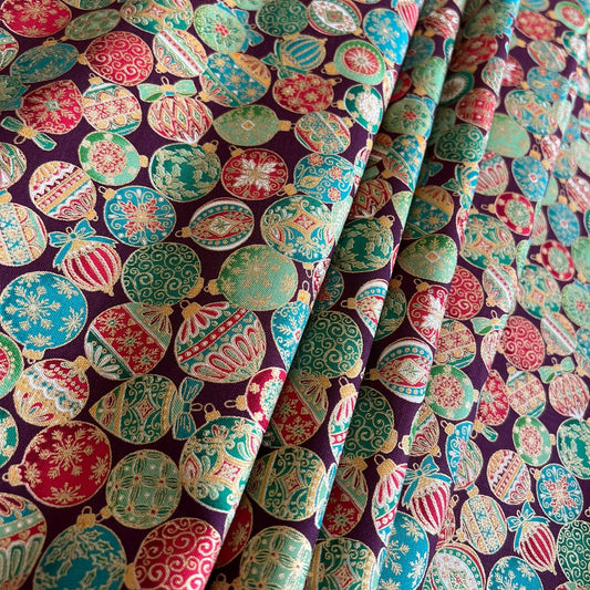 A close-up of patterned fabric with a repeating Christmas ornament print on a dark burgundy background. The ornaments are in green, red, pink, and metallic gold, with some featuring snowflakes and striped details.