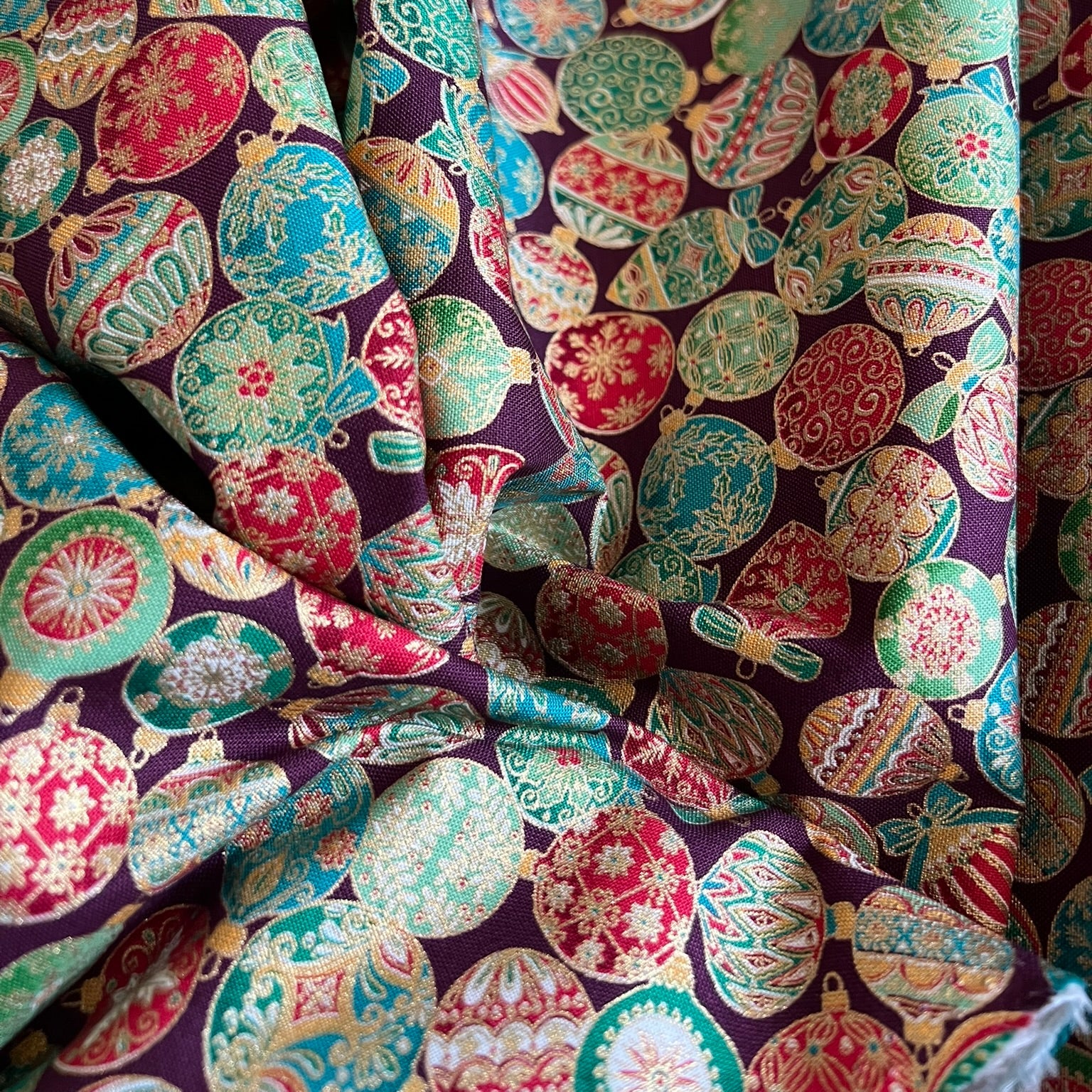 A close-up of draped fabric featuring a festive Christmas ornament pattern on a dark purple background, with green, red, green, and metallic gold ornaments.