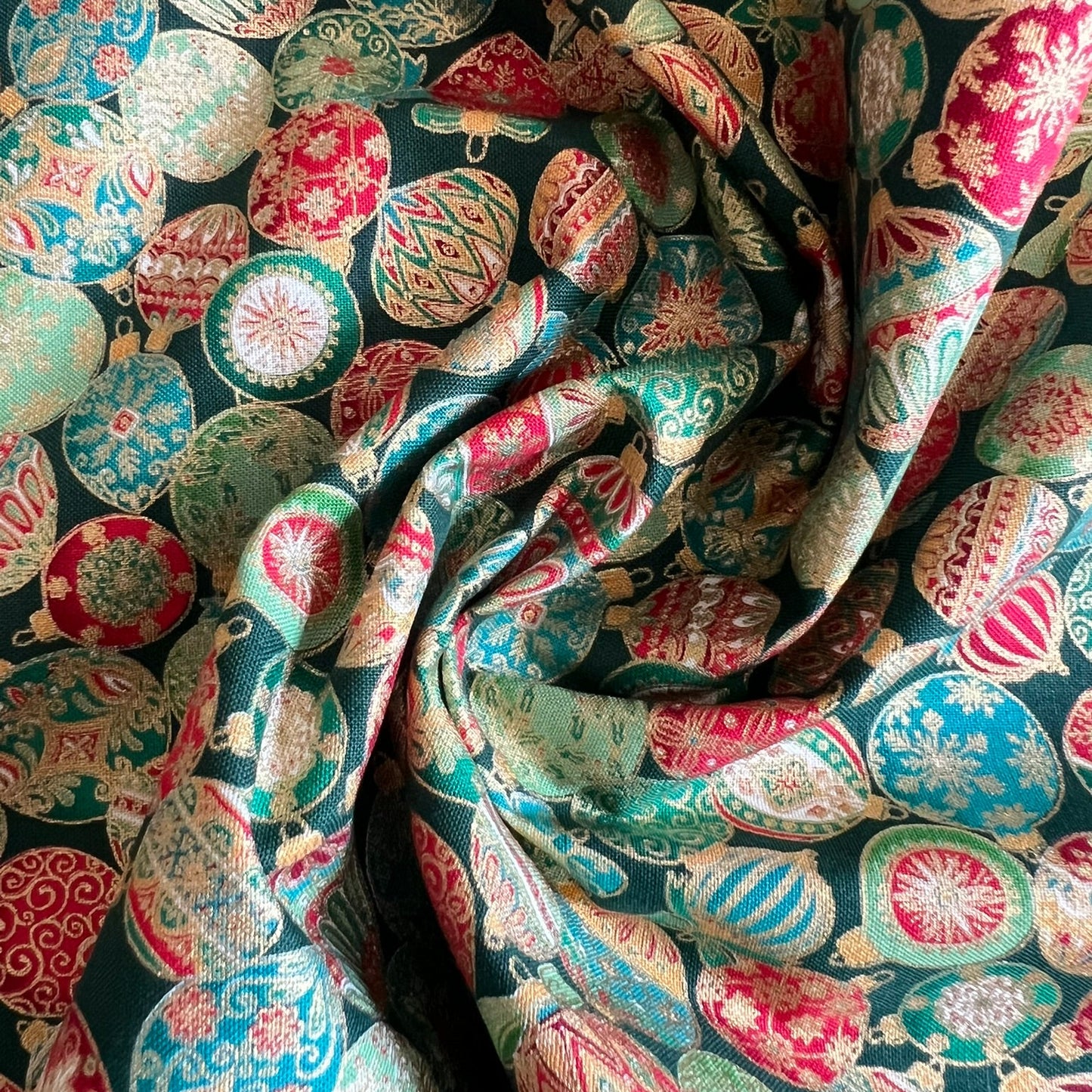 A close-up of draped fabric with an all-over Christmas ornament pattern in green, red, cream, and metallic gold on a dark background.