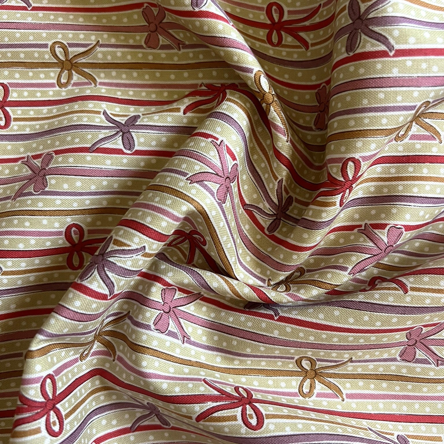 A close-up of patterned fabric with horizontal white stripes, thin red and mauve lines, small white dots, and repeating bow motifs in red, mauve, and tan.