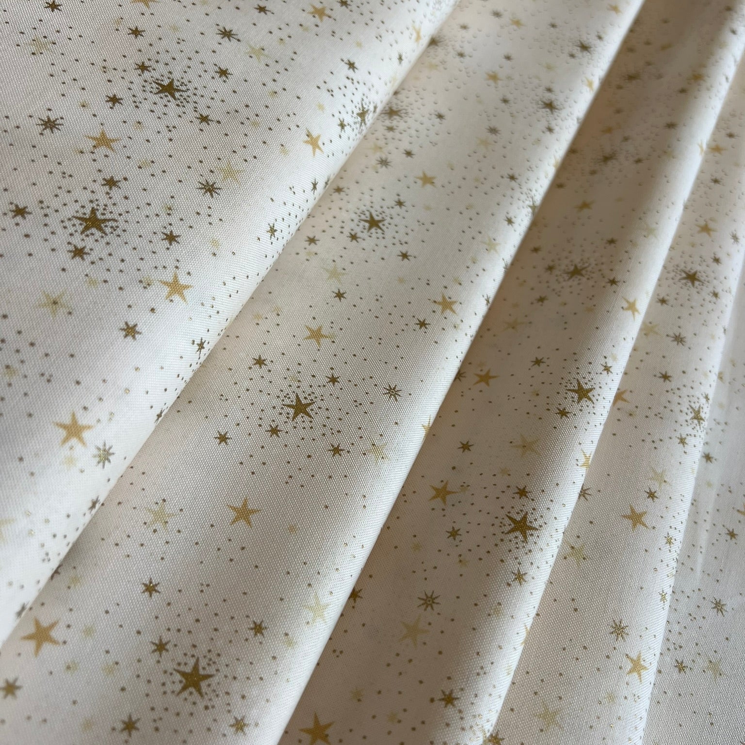 A close-up of cream-colored fabric with a scattered gold star and dot pattern, arranged in soft diagonal folds.