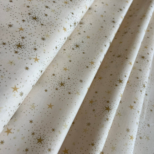 A close-up of cream-colored fabric with a scattered gold star and dot pattern, arranged in soft diagonal folds.