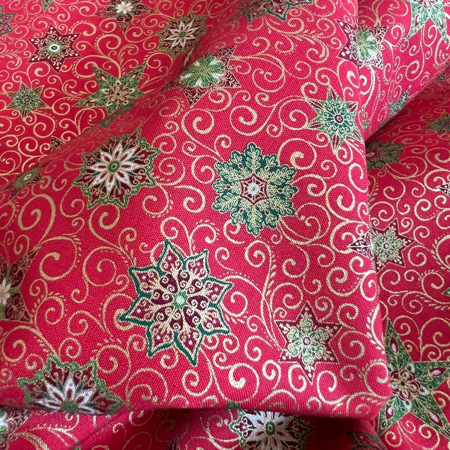 A close-up of a red patterned fabric with green, metallic gold and beige floral medallions and swirling motifs, draped in soft folds.