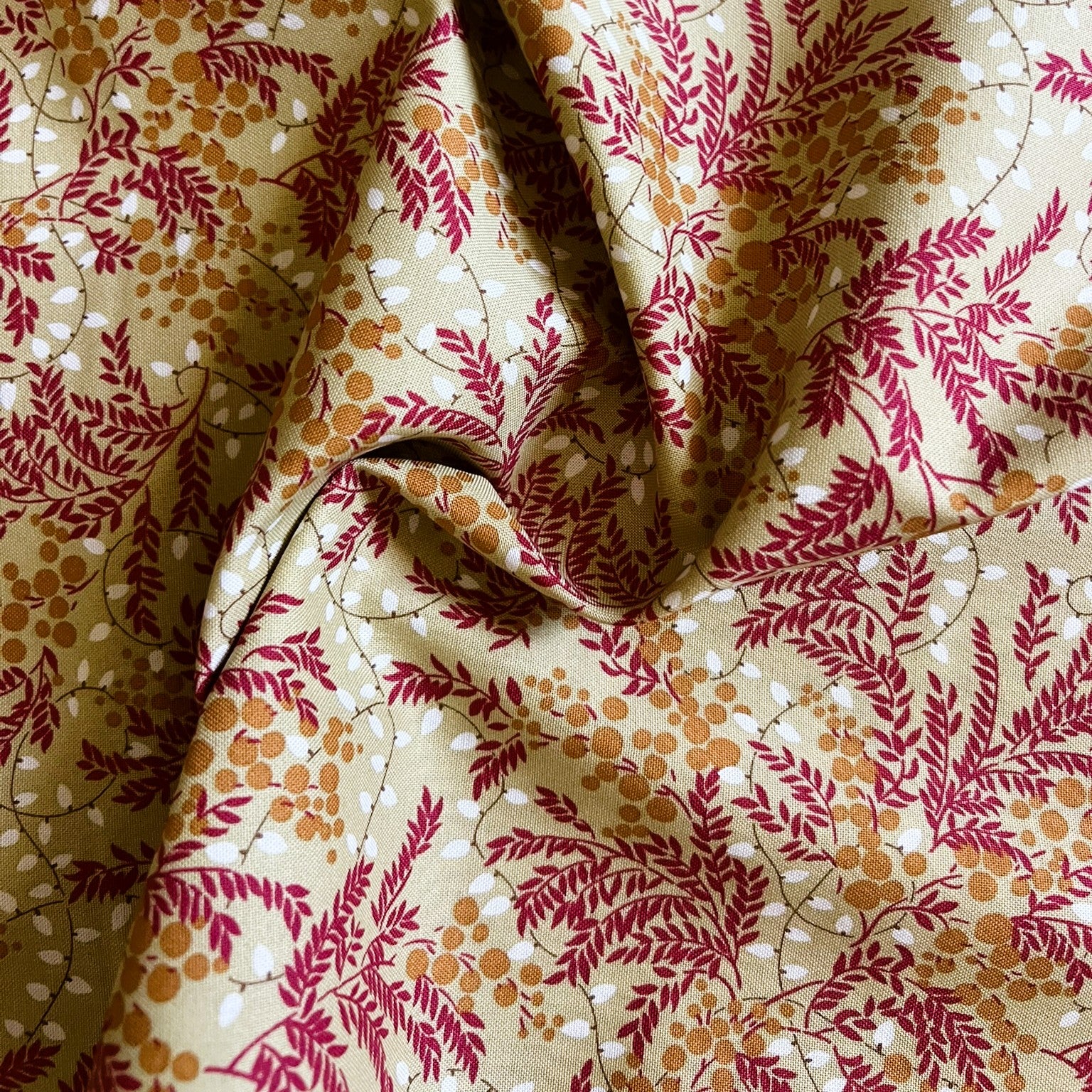 A close-up of draped fabric with an allover botanical print featuring dark red leafy vines, small orange-brown dots, and thin dark gray stems on a warm cream background.